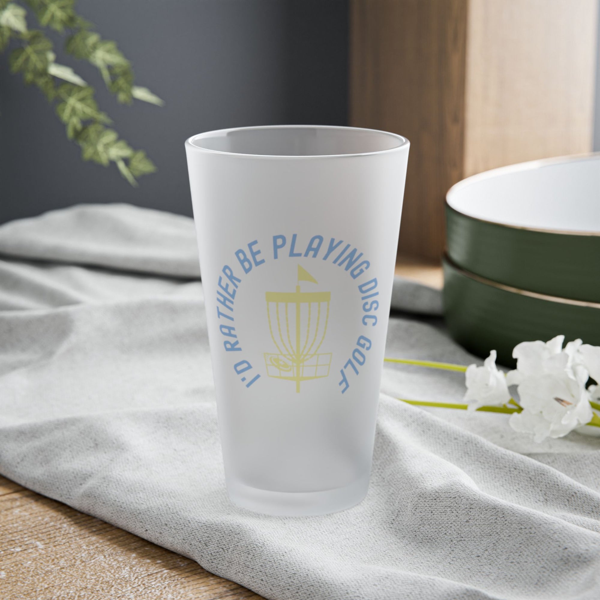 A frosted pint glass with disc golf chains and basket with the text "I'd Rather Be Playing Disc Golf" surrounding them, on a tabletop