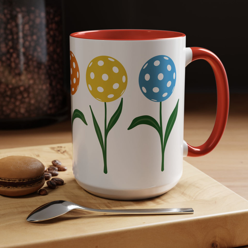 A ceramic coffee mug with red accents on the handle and interior, with colorful flowers made from pickleballs on green stems with leaves, in a coffee house setting