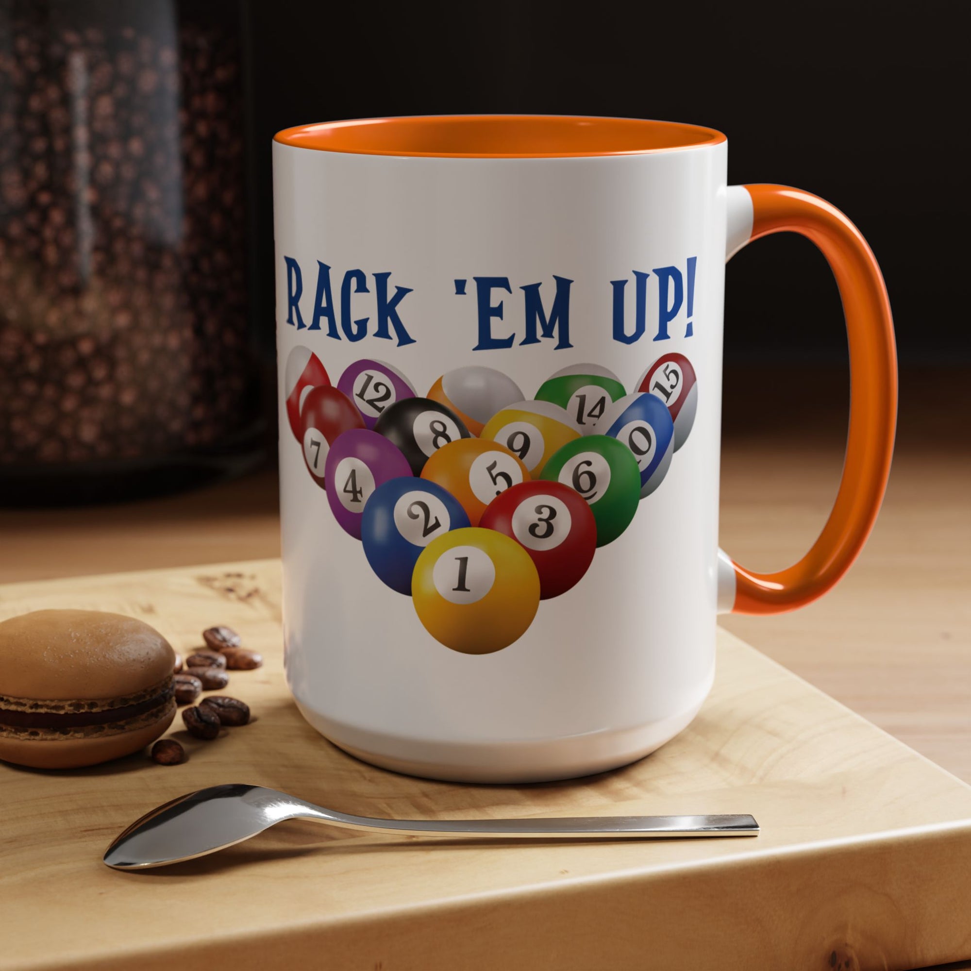 A white ceramic coffee mug with orange accents on the handle and interior, with racked pool ball design and 'Rack 'Em Up!' text, in a coffee shop setting
