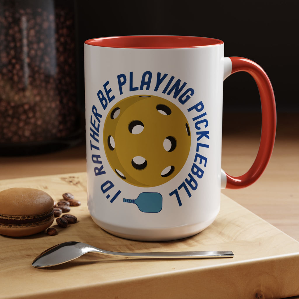 Coffee mug with red accents on the handle and inside, with a yellow pickleball and blue pickleball paddle with the words "I'd Rather Be Playing Pickleball" set in a coffeeshop setting.