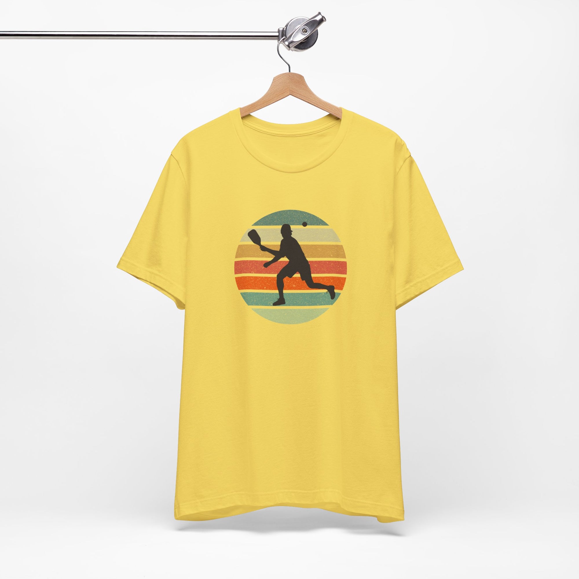 Yellow t-shirt with a graphic design of a person playing pickleball on a hanger against a white background