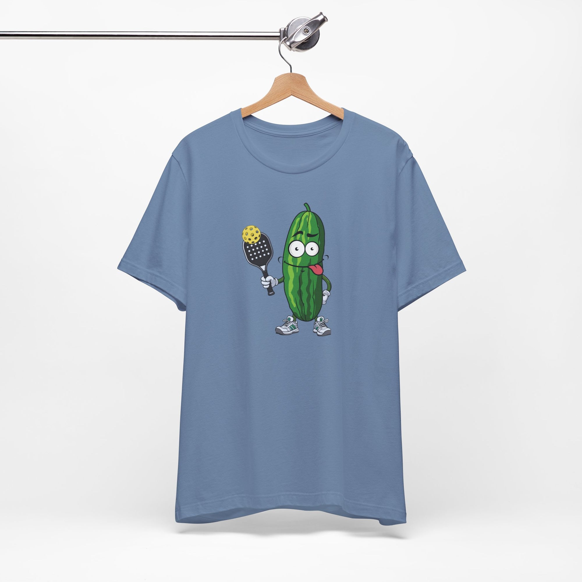 Steel blue t-shirt with a cartoon pickle with googly eyes and his tongue sticking out, holding a pickleball paddle with a yellow pickleball, on a hanger against a white background
