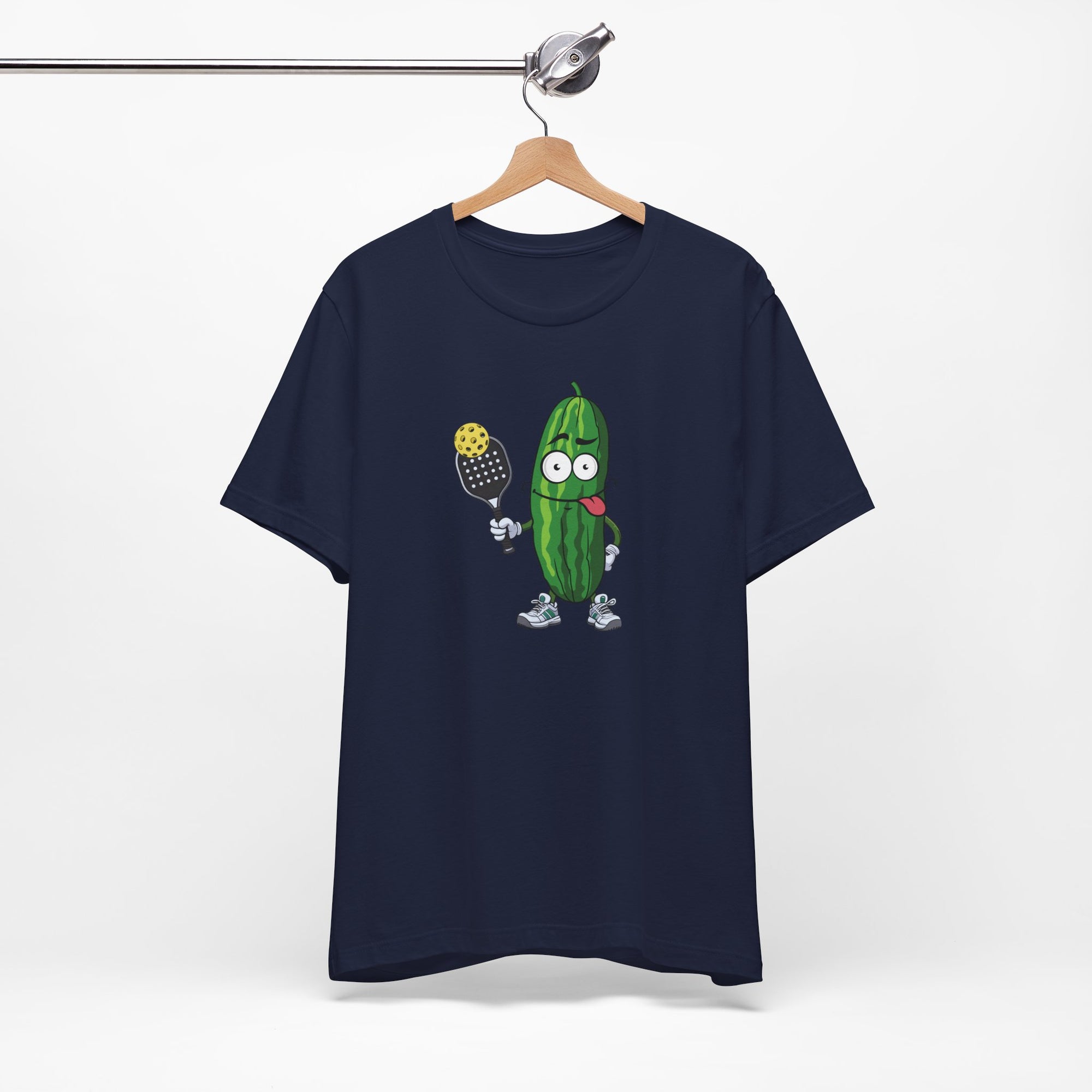 Navy blue t-shirt with a cartoon pickle with googly eyes and his tongue sticking out, holding a pickleball paddle with a yellow pickleball, on a hanger against a white background