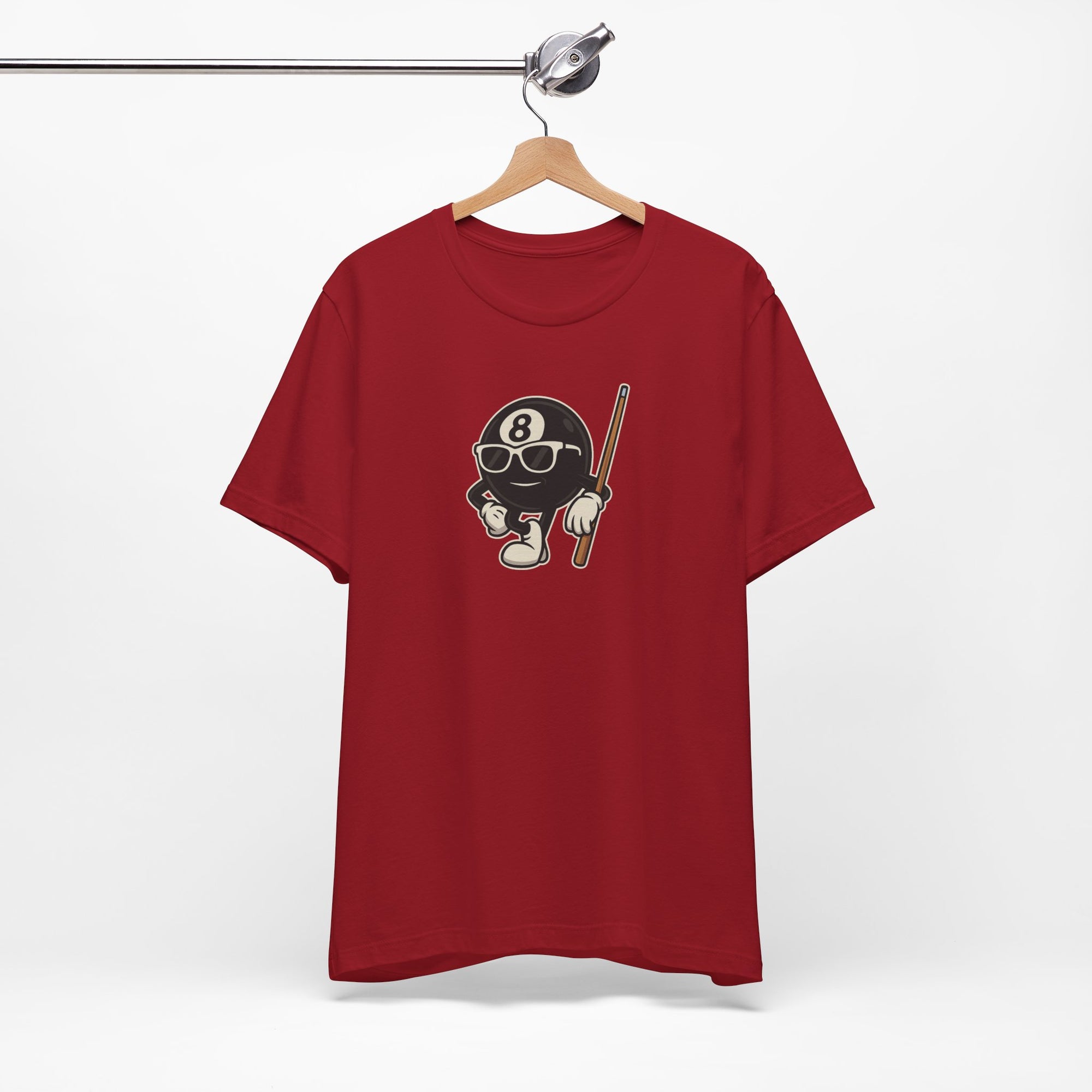 Red t-shirt with a graphic of a cartoon billiards 8 ball with arms and legs and wearing sunglasses and leaning on a cue stick, on a hanger against a white background