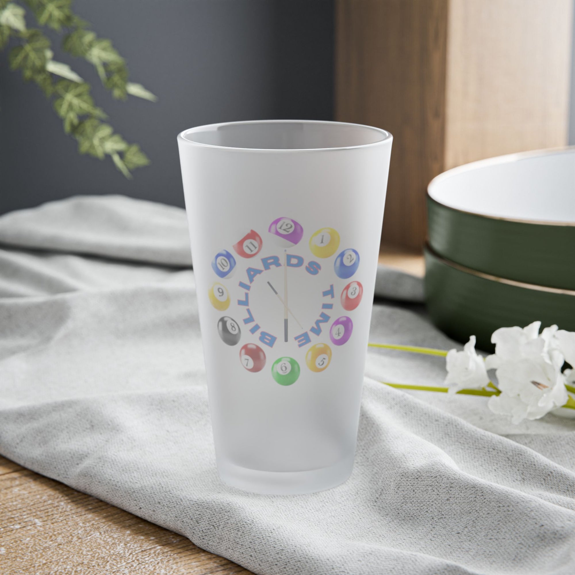 A frosted pint glass with the text "Billiards Time" surrounded by a clock made up of colorful billiard balls and cue sticks, on a tabletop