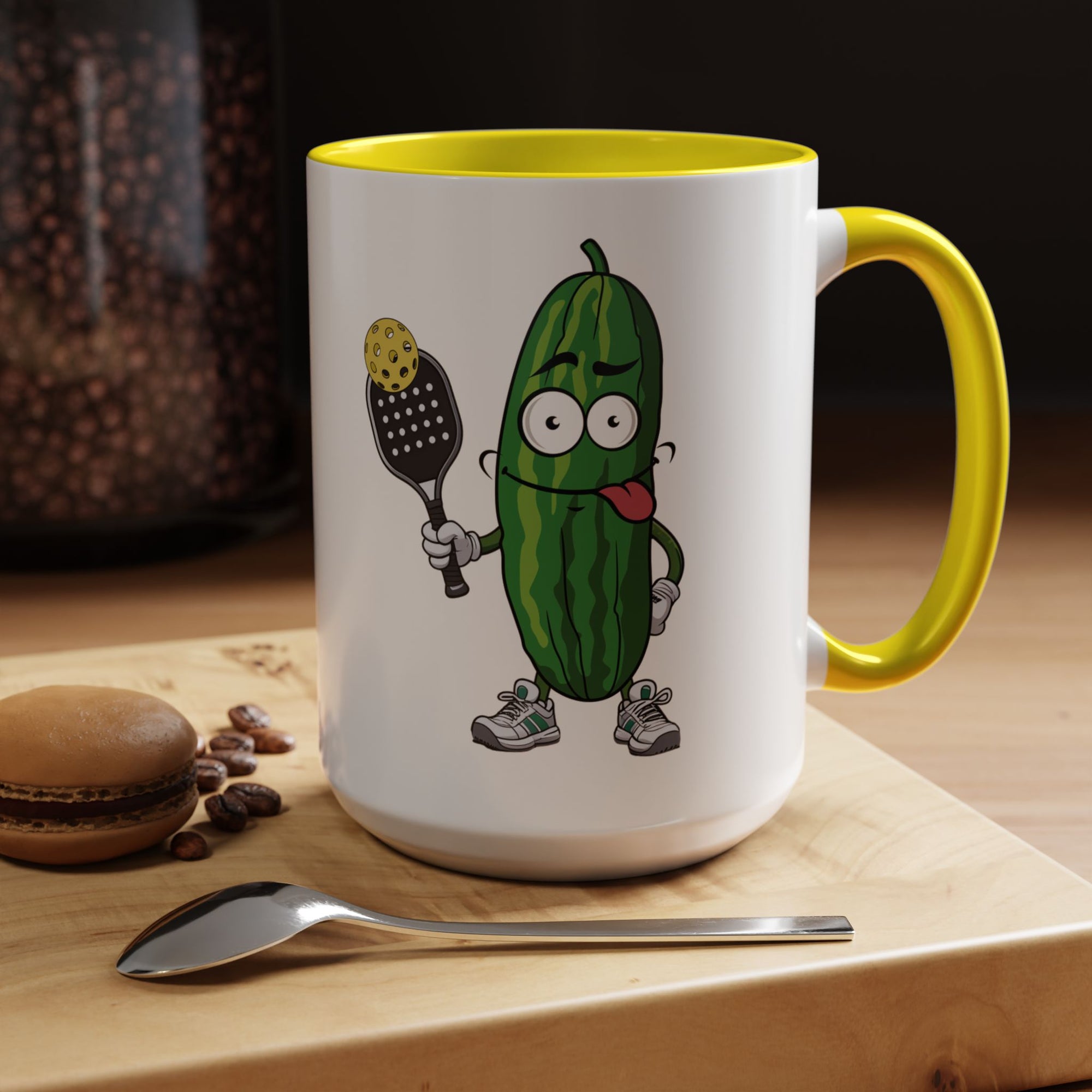 A ceramic coffee mug with yellow accents on the handle and interior, with a cartoon pickle with googly eyes and his tongue sticking out, holding a pickleball paddle with a yellow pickleball, in a coffee shop setting