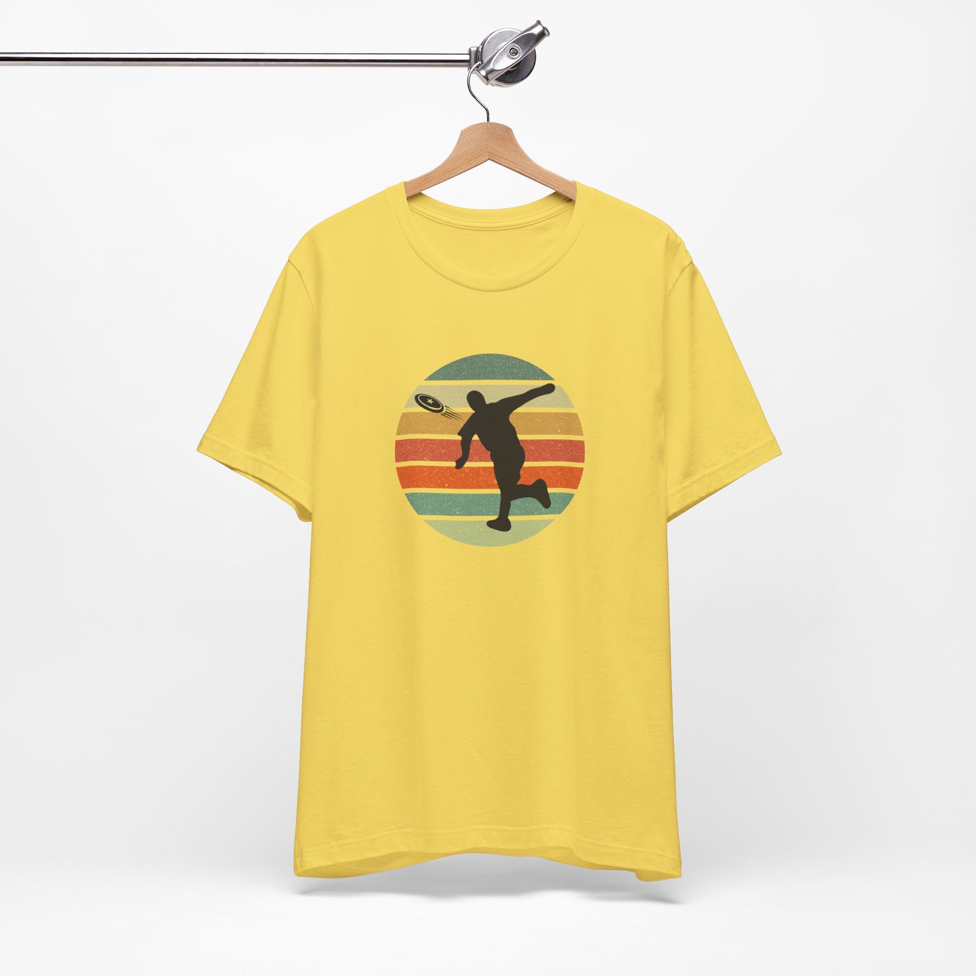 Yellow t-shirt with a graphic of a silhouette disc golf player throwing a disc above a colorful retro graphic, hanging on a wooden hanger.