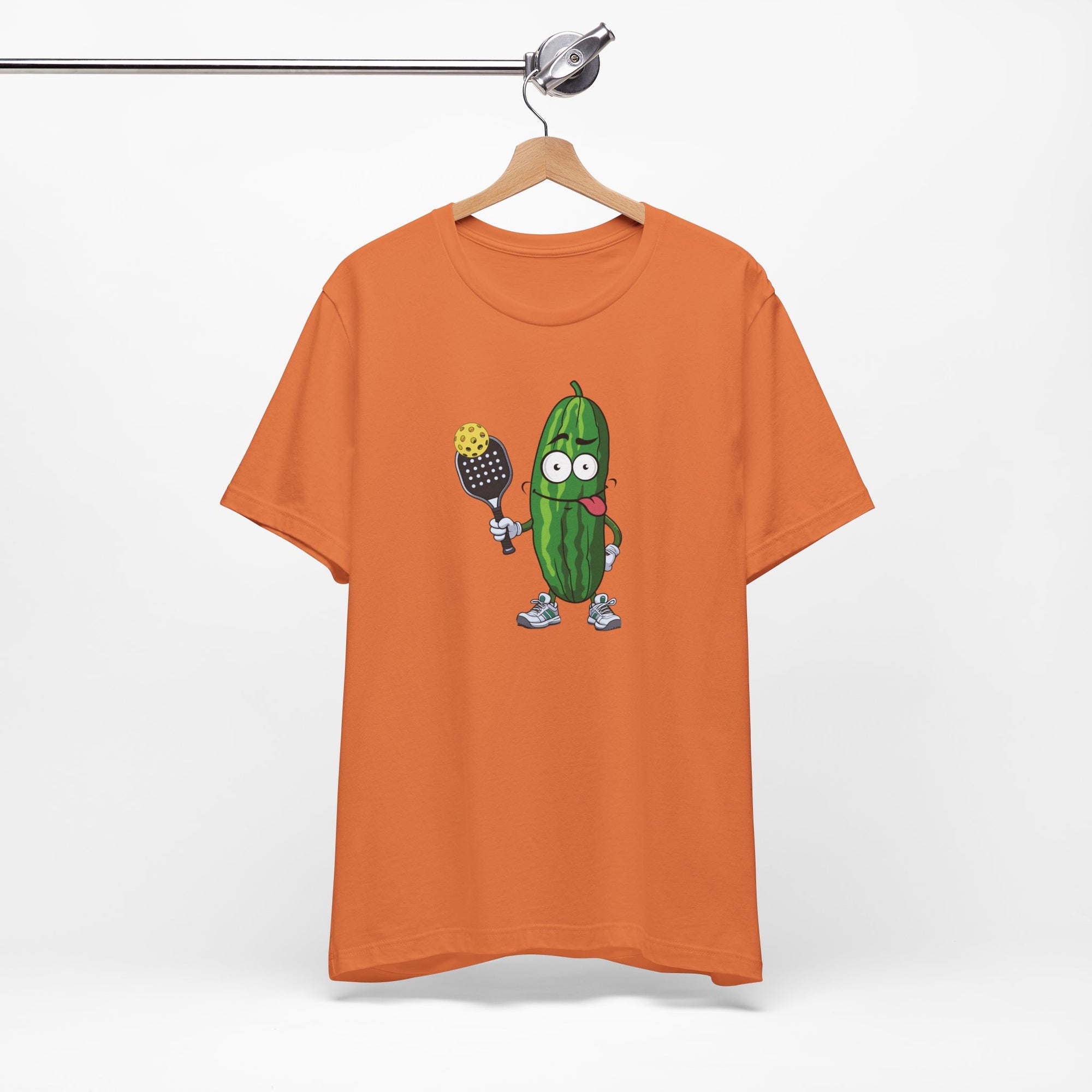 Orange t-shirt with a cartoon pickle with googly eyes and his tongue sticking out, holding a pickleball paddle with a yellow pickleball, on a hanger against a white background